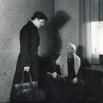 A person in a black coat carrying a bag speaking with and looking down at an elderly woman