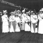A group of women dressed in white, holding flags and items celebrating suffrage